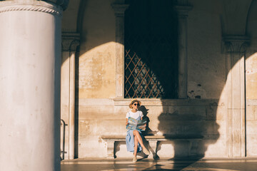 Cheerful young female tourist holding map and sitting on bench at building