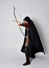 Full length portrait of girl with red hair wearing medieval archer costume with black cloak. Standing pose with back to the camera holding a bow and arrow,  isolated against a grey studio background.