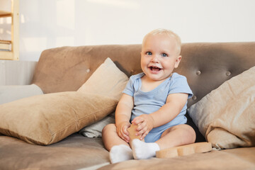 Horizontal portrait shot of cute little baby boy wearing light blue clothes sitting on sofa smiling at camera, copy space