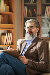 Vertical medium shot of handsome bearded man wearing fashionable outfit sitting on sofa in modern office room making notes