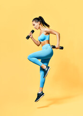 Fototapeta na wymiar Sporty woman in sportswear jumping with dumbbells on yellow background