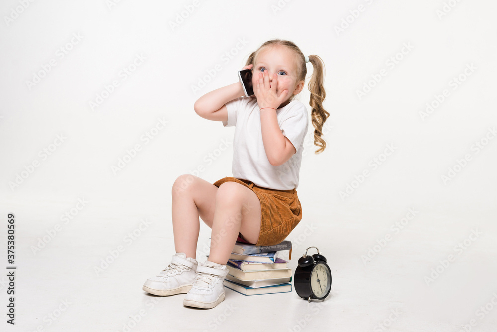 Wall mural Portrait of a little girl talk phone sitting on a stack of books over white background.