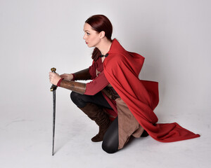 Full length portrait of girl with red hair wearing medieval costume and red cloak. Sitting pose, ...