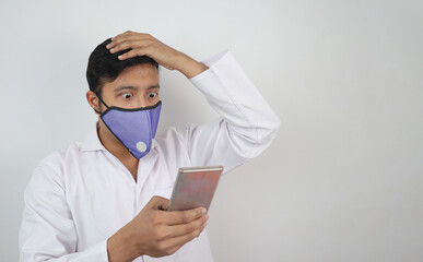 a male doctor with mask and white coat shocked to see a news in his mobile phone in hand isolated in white grey background with copy space for text.