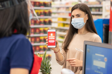 Modern woman wearing mask on face using digital discount coupon on her smartphone in supermarket - obrazy, fototapety, plakaty