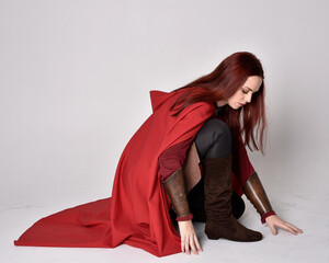 Full length portrait of girl with red hair wearing medieval costume and red cloak. Sitting pose, ...