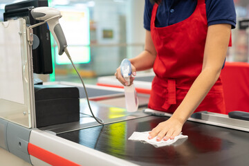 Unrecognizable cashier wearing red apron cleansing checkout surface with disinfectant in modern...