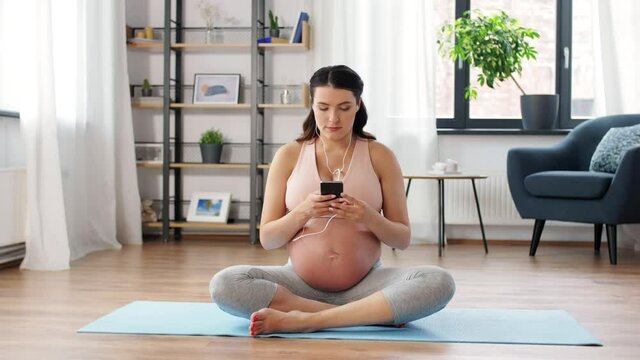 pregnancy, sport and people concept - happy pregnant woman with earphones and smartphone doing yoga at home