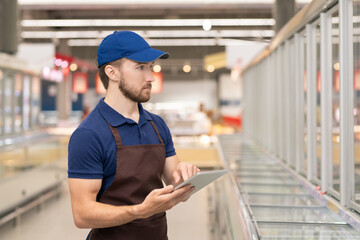 Young man wearing uniform working as merchandiser in modern supermarket using digital tablet,...