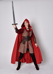 Full length portrait of girl wearing medieval costume and red cloak. Standing pose holding a sword,  isolated against a grey studio background.