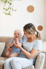 Vertical shot of Caucasian woman sitting on sofa having great time with her cheerful little baby, copy space