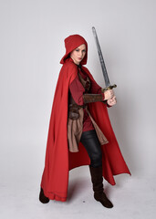 Full length portrait of girl wearing medieval costume and red cloak. Standing pose holding a sword,  isolated against a grey studio background.