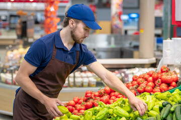 Modern supermarket worker wearing uniform setting out fresh vegetables, horizontal medium shot, copy space