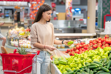 Portrait of young adult woman standing in supermarket with shopping cart choosing sweet peppers