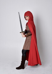 Full length portrait of girl wearing medieval costume and red cloak. Standing pose holding a sword,  isolated against a grey studio background.