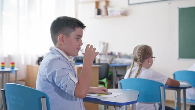 schoolboy drinking water while sitting at a desk in the classroom during a lesson on background of classmates, elementary school
