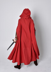Full length portrait of girl wearing medieval costume and red cloak. Standing pose holding a sword,...