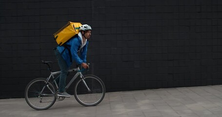Side view of tired male courier with food bag riding bicycle and wiping forehead at street. Young man in protective helmet delivering order to custumer. Concept of food delivery