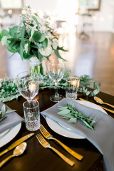 A beautiful table setting at a wedding with wine glasses, votive candles, a bridal bouquet, gold silverware, and eucalyptus leaves. 