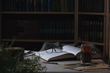 Eyeglasses lying on the opened book and a cup of tea. Bookcases in the background