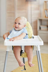 Vertical portrait of cute little child sitting on high chair waiting for food looking at something, copy space