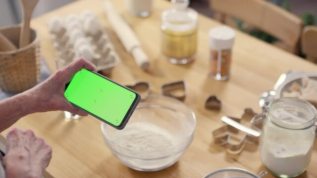High angle shot of unrecognizable senior woman using mobile phone with green screen on it. Baking ingredients and tools on kitchen table