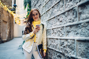 Portrait of happy female blogger with education equipment in hand standing near wall and smiling during mobility time for online socialising, attractive red hair hipster girl holding smartphone gadget