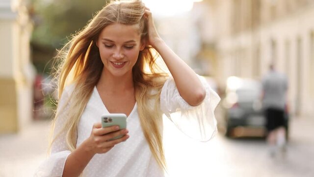 Portrait of attractive blonde woman texting message on smartphone on city street in slow motion