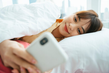 Obraz na płótnie Canvas Asian dark hair woman laying on a bed using smartphone in the morning.