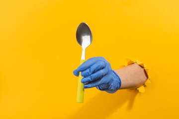 Hand in a latex glove giving a metal spoon through torn yellow wall