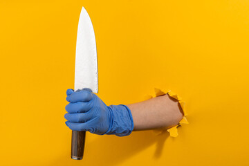 Hand in a latex glove holding a Japanese knife through torn yellow wall