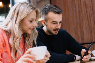 Smiling caucasian female and male colleagues spending coffee break together watching vlog on smartphone, positive hipster guys searching information in social media using mobile phone and 4G