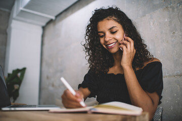 Positive curly dark skinned woman writing in notepad and talking on mobile phone in cafe, cheerful african american 20s female blogger writing article and communicate via smartphone smiling