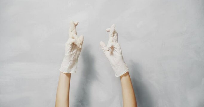Close up of female hands in protective medical gloves showing crossed fingers on gray background.