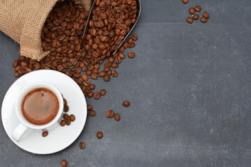 Black coffee cup with foam, beans in scoop scattered from burlap bag on dark table background with copy space. Arabica grains. Coffee shop, caffeine, roast concept