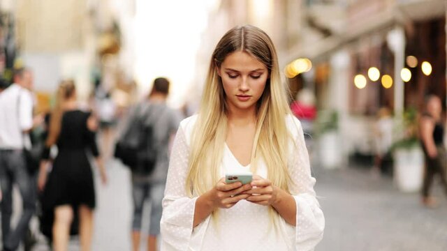 Beautiful blonde woman in a white dress looking up at street signs and map trying to find her way using her mobile phone. Girl using map application outdoors