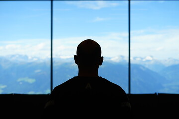 Silouette of a bald man watching the mountain from the window of an alpine refuge - obrazy, fototapety, plakaty