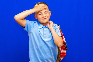 scared little boy with open mouth, in blue t shirt striped backpack keeping hands near head blue background