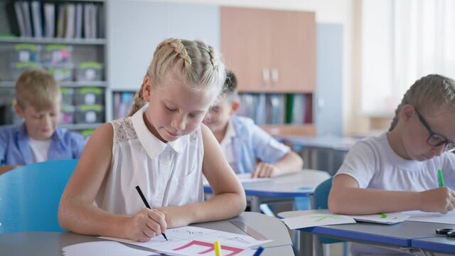 portrait of happy pupil girl doing classwork sitting at a desk on background of classmates at elementary school