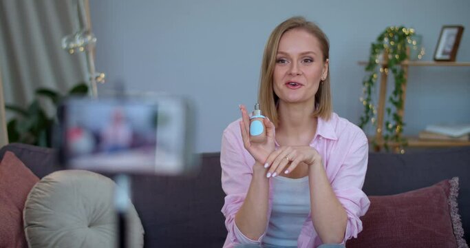 Blonde woman vloger making video review about make up product while sitting on couch at home.Cheerful girl talking and showing skin foundation while recording herself on smartphone.