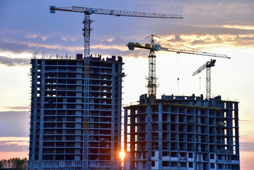 Tower cranes in action at construction site on sunset background. Crane the build the high-rise building. New residential skyscraper. Tall house renovation project, government programs