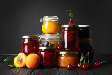 Jars with different jams and fresh fruits on black wooden table
