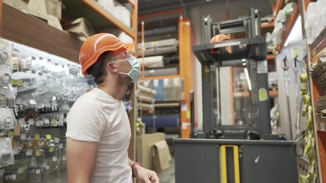 Caucasian industrial worker in orange hard hat, safety goggles and medical mask in hardware store against the background of a loading cargo lift. Male warehouse worker mask coronavirus, covid 19
