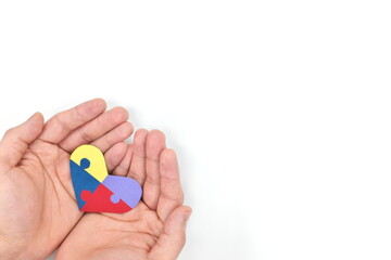 Cupped male hands holding a colorful puzzle heart shape paper cutout. Top view flat lay in white background with copy space. World Autism awareness day, support and care concept.