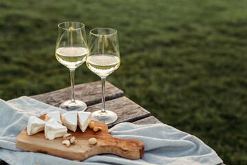 Two glasses of white wine and wooden plate with cheese and nuts during sunset time outside. - obrazy, fototapety, plakaty