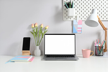 A white blank screen laptop is putting on a white working desk surrounded by various equipment.