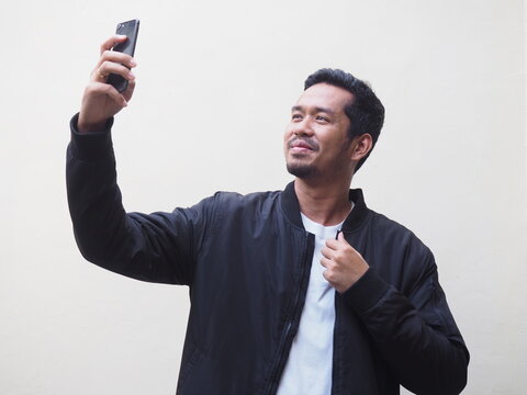 A handsome adult Asian man wearing black bomber jacket and white shirt taking a selfie photo using mobile phone isolated on white. Space for copy 