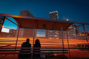 An illuminated city street at the night. The bus stop with silhouettes of a couple, sitting on the bench, in front of bright light trails, from moving cars.