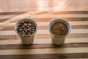 On a wooden plank there are two cups filled with whole roasted coffee beans and instant coffee powder.
