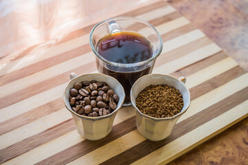 There are three cups of coffee on a wooden board, one with roasted black coffee beans, the second with brewed coffee, and the third with instant coffee.